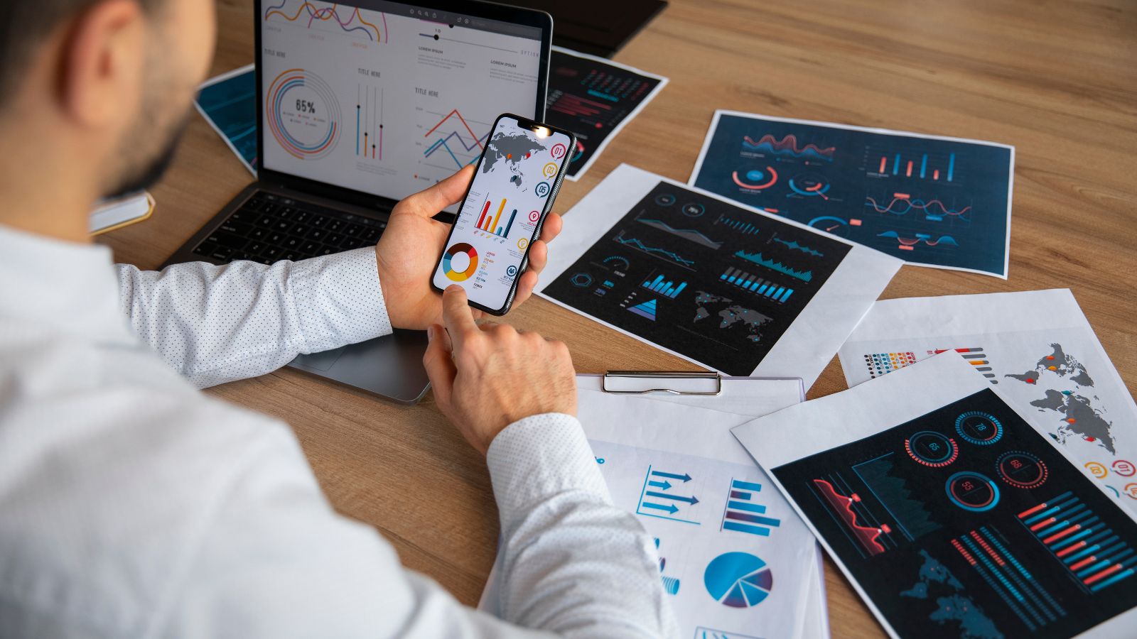 Um profissional analisa gráficos e indicadores em seu smartphone enquanto compara relatórios impressos e dashboards no notebook sobre a mesa, aplicando a análise de dados para negócios na prática.